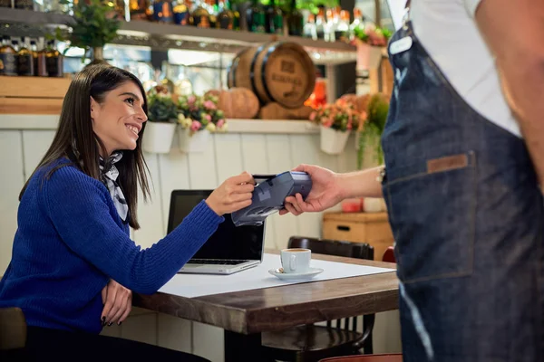 Smiling attractive caucasian brunette with long hair sitting in cafe and paying bill with credit card. Waiter holding POS terminal and standing next to woman. — Stock Photo, Image