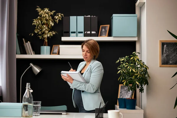 Czarująca, biała, ciężarna bizneswoman w garniturze, stojąca w biurze i robiąca listę w swoim notesie. Nowoczesne wnętrze biura. — Zdjęcie stockowe
