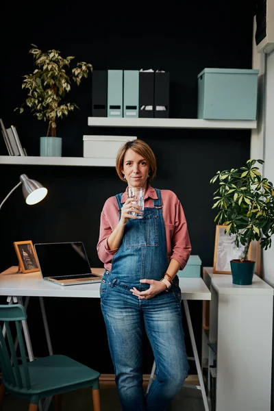 Bájos pozitív kaukázusi terhes nő áll otthon iroda, támaszkodik az asztalra, kezében pohár víz és megható has. — Stock Fotó