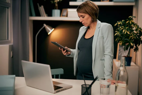 Modern ofisinde duran, e-posta okumak ve masaya yaslanmak için tablet kullanan takım elbiseli, çalışkan hamile bir iş kadınına odaklanmış. Gece işi konsepti. — Stok fotoğraf