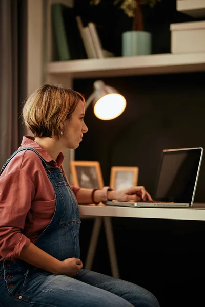 Ciddi bir beyaz hamile esmer ofiste oturuyor, iş için dizüstü bilgisayar kullanıyor ve karnına dokunuyor.. — Stok fotoğraf