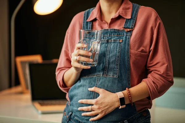 Close up of pregnant caucasian woman leaning on the desk in her home office, touching belly and holding glass of water. — Stock Photo, Image