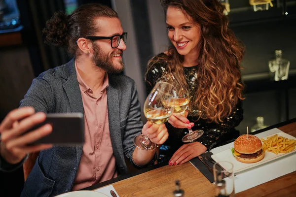 Mignon caucasien à la mode couple assis dans restaurant au dîner tenant des verres de vin et de prendre selfie . — Photo