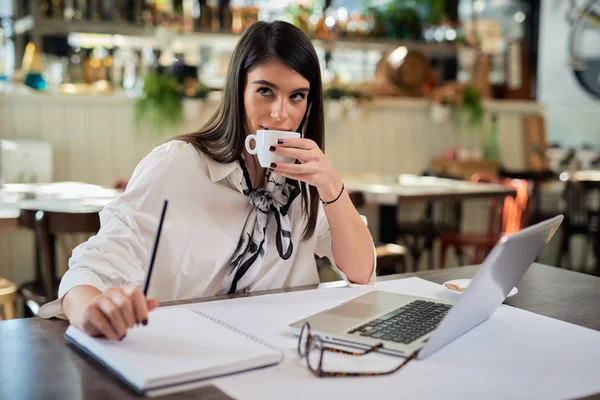 Atraktivní kavkazská elegantní brunetka sedící v kavárně, popíjející kávu a dívající se do kamery. Na stole je notebook. — Stock fotografie