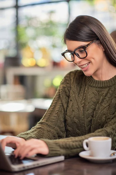 微笑积极的高加索黑发与眼镜穿着休闲坐在咖啡店和做她的自由职业工作在笔记本电脑。 手放在键盘上. — 图库照片
