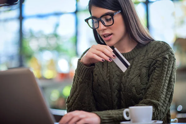 Atrakcyjna biała brunetka w swetrze siedząca w kawiarni, pisząca na klawiaturze i trzymająca kartę kredytową. — Zdjęcie stockowe