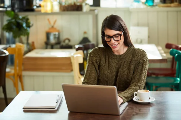 Sorrindo morena caucasiana positiva com óculos vestidos casuais sentados no café e fazendo seu trabalho freelance no laptop. As mãos estão no teclado . — Fotografia de Stock
