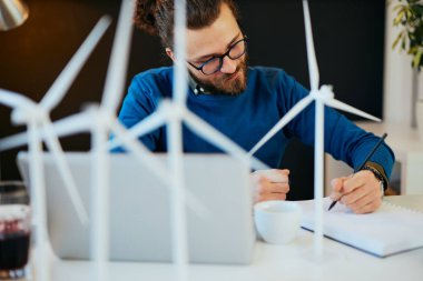 Genç, yenilikçi, kendini işine adamış sakallı bir çalışan modern ofisinde oturup yel değirmenlerinin taslaklarını çiziyor. Sürdürülebilir geliştirme kavramı.