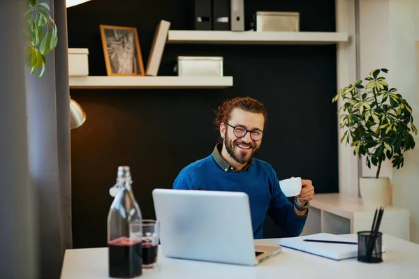 Joven sonriente guapo caucásico freelancer sentado en su moderna oficina en casa y beber café . — Foto de Stock