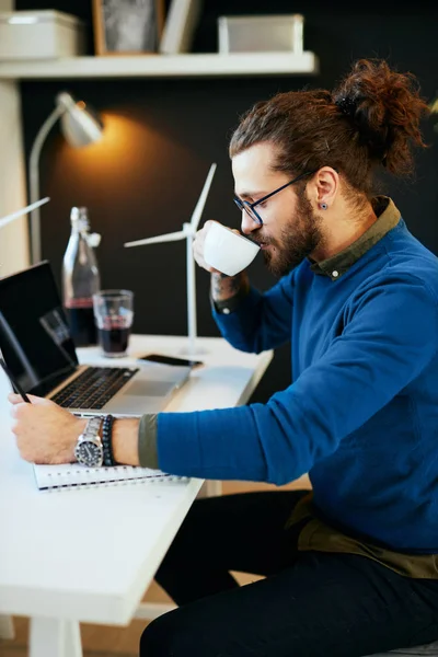 Νέος καινοτόμος, αφοσιωμένος γενειοφόρος εργαζόμενος κάθεται στο σύγχρονο γραφείο του και σχεδιάζει σκίτσα ανεμόμυλων και πίνοντας καφέ. Έννοια της αειφόρου ανάπτυξης του εργαζομένου που κάθεται στη θέση του — Φωτογραφία Αρχείου