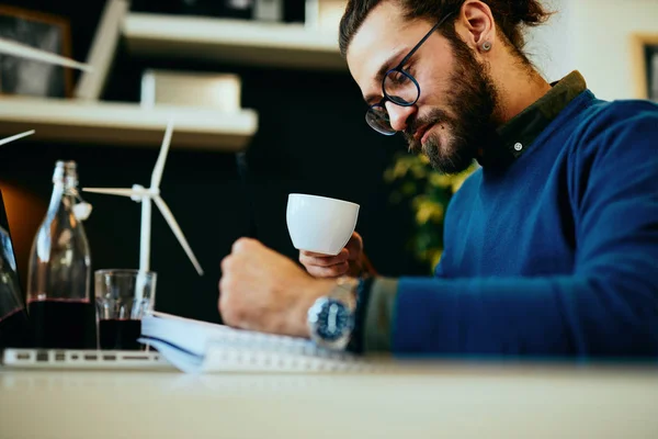 Νέος καινοτόμος, αφοσιωμένος γενειοφόρος εργαζόμενος κάθεται στο σύγχρονο γραφείο του και σχεδιάζει σκίτσα ανεμόμυλων και πίνοντας καφέ. Έννοια της αειφόρου ανάπτυξης. — Φωτογραφία Αρχείου