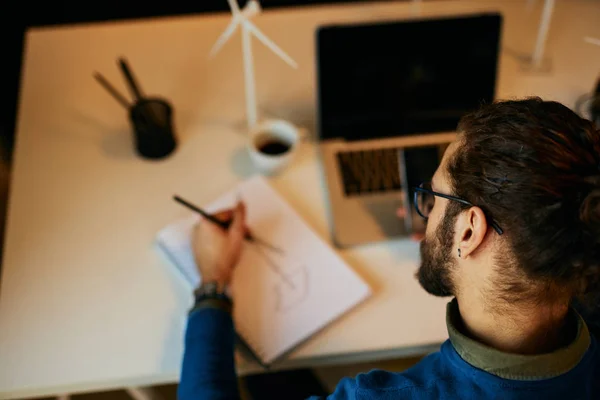 若い革新的な献身的な髭の従業員は、彼のモダンなオフィスに座って、風車のスケッチを描きます。持続可能な開発の概念. — ストック写真