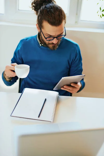 Jeune coûteux attrayant caucasien barbu pigiste assis dans son bureau à la maison, en utilisant une tablette pour lire les nouvelles et tenant une tasse de café . — Photo