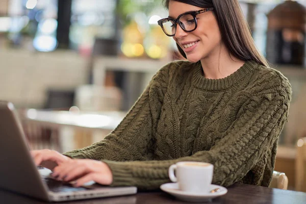 Usmívající se pozitivní kavkazské brunetky s brýlemi oblečené ležérně sedí v kavárně a dělá svou práci na volné noze na notebooku. Ruce jsou na klávesnici. — Stock fotografie