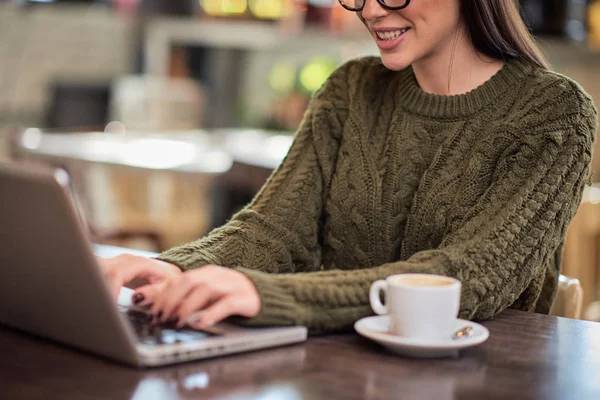 Usmívající se pozitivní kavkazské brunetky s brýlemi oblečené ležérně sedí v kavárně a dělá svou práci na volné noze na notebooku. Ruce jsou na klávesnici. — Stock fotografie