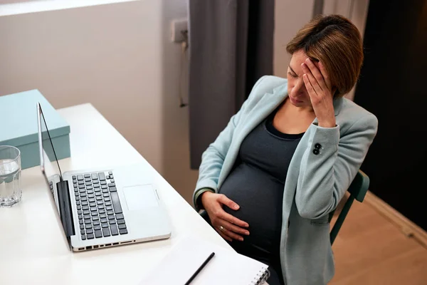 40 'lı yaşlarda hamile bir iş kadını ofisinde oturmuş karnına dokunuyor ve baş ağrısı çekiyor.. — Stok fotoğraf