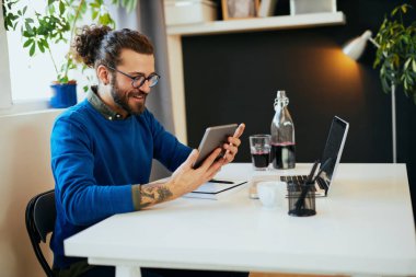 Genç, gülümseyen, beyaz sakallı, grafik tasarımcı ofisinde oturuyor ve tablet kullanıyor..