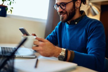 Genç, sakallı hippi evinde oturmuş elinde bir fincan kahveyle akıllı telefon kullanıyor..