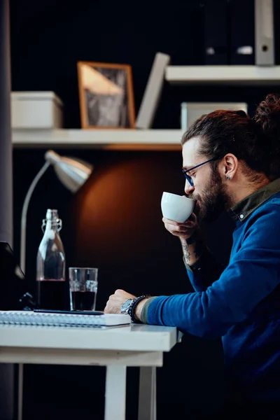 Sidovy över attraktiv skäggig entreprenör sitter på sitt kontor, koppla av och dricka kaffe. — Stockfoto