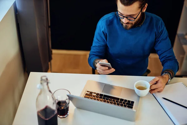 Joven guapo barbudo hipster sentado en su oficina en casa, sosteniendo una taza de café y usando un teléfono inteligente. — Foto de Stock