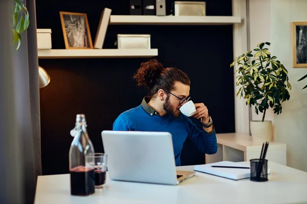 Ung attraktiv kaukasisk skäggig frilansare sitter på sitt hemmakontor och dricker morgonkaffe. På bordet är laptop. — Stockfoto