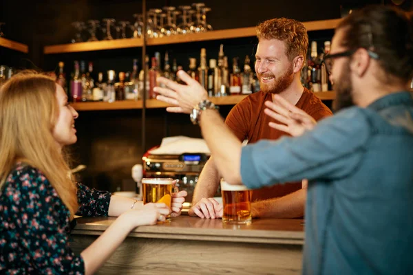 Vrolijke vrienden leunend op een bar, bier drinkend en babbelend met de barman. Avond uit.. — Stockfoto