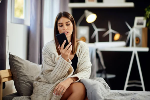 Hermosa morena caucásica sonriente sentada vestida con suéter beige sentada en la cama en el dormitorio y usando un teléfono inteligente . — Foto de Stock