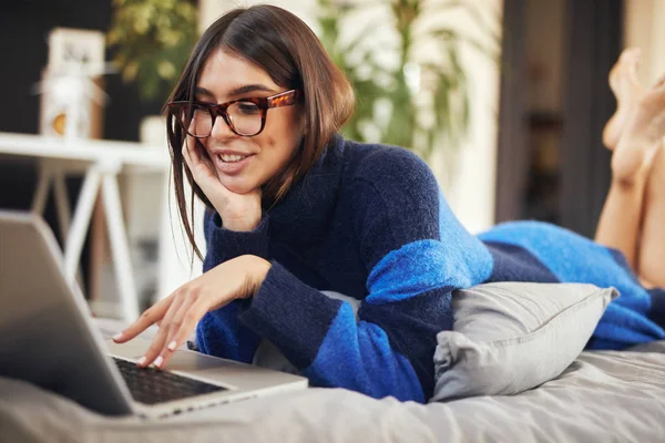 Attractive caucasian brunette in blue and black sweater lying on stomach in bed and typing on laptop. — Stock Photo, Image