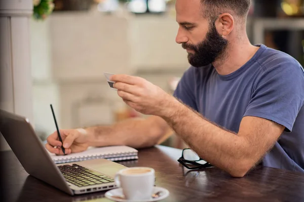 Guapo barbudo caucásico hombre sosteniendo tarjeta de crédito y anotando gastos mientras está sentado en la cafetería . — Foto de Stock