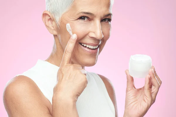 Gorgeous smiling Caucasian senior woman trying out new anti age cream and looking at camera. Beauty photography. — Stock Photo, Image