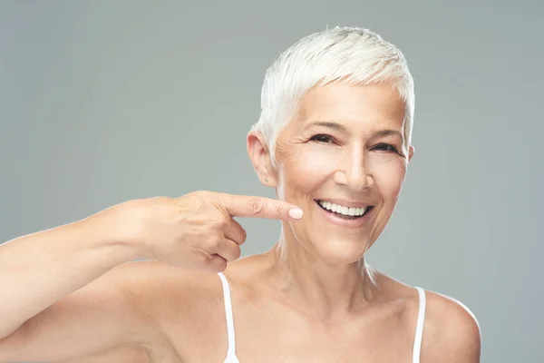 Hermosa mujer mayor caucásica sonriente con el pelo gris corto apuntando a sus dientes y mirando a la cámara. Belleza fotografía . — Foto de Stock