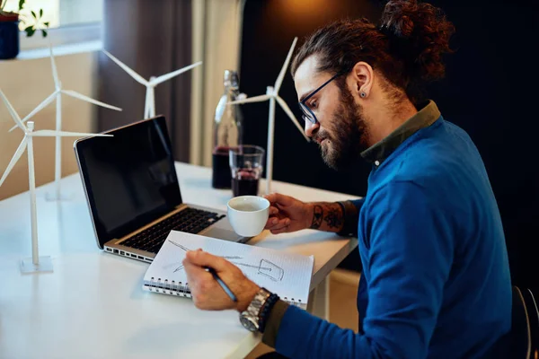 Primer plano del ingeniero caucásico innovador dedicado sentado en su oficina y dibujando molino de viento. Concepto de desarrollo sostenible . — Foto de Stock