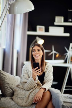 Güzel, gülümseyen, beyaz tenli esmer, bej bir süveter giymiş yatak odasında oturuyor ve akıllı telefon kullanıyor..