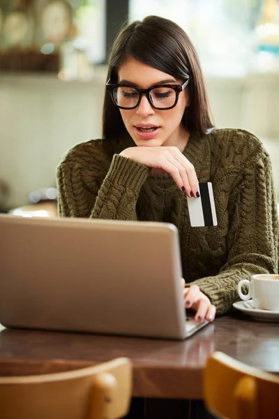 Urocza biała brunetka siedzi w kawiarni, trzyma kartę kredytową i używa laptopa do zakupów online. — Zdjęcie stockowe