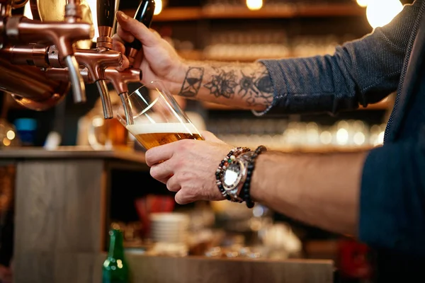 Καυκάσιος μπάρμαν με τατουάζ που σερβίρει μπύρα ενώ στέκεται στην παμπ. Επιλεκτική εστίαση στο χέρι. — Φωτογραφία Αρχείου