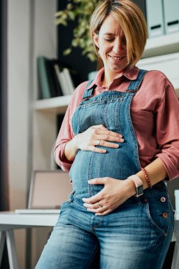 Gururlu, beyaz, müstakbel anne ev ofisinin masasına yaslanıp kameraya bakarken karnına dokunuyor..