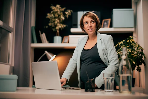 Glimlachende vrolijke blanke zwangere zakenvrouw in pak staan in haar moderne kantoor en met behulp van laptop. — Stockfoto