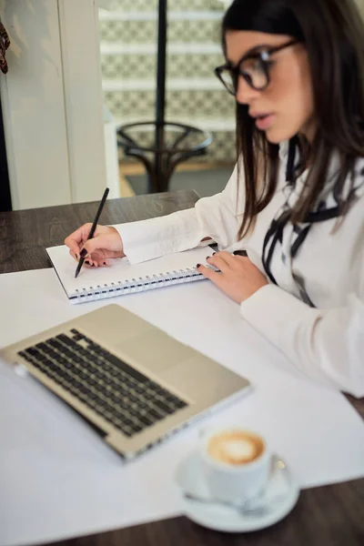 Pozitivní atraktivní mladá podnikatelka ve třicátých letech oblečená v košili sedí v kavárně a psaní úkolů v agendě. Na stole je laptop. Vzdálený obchodní koncept. — Stock fotografie