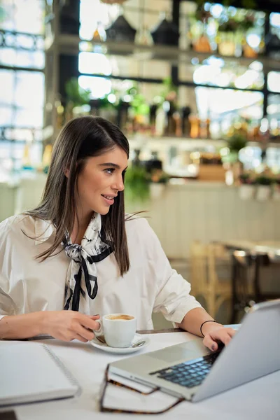 Aantrekkelijke Kaukasische elegante brunette zittend in cafe, koffie drinkend en kijkend naar laptop. Op tafel ligt een laptop. — Stockfoto