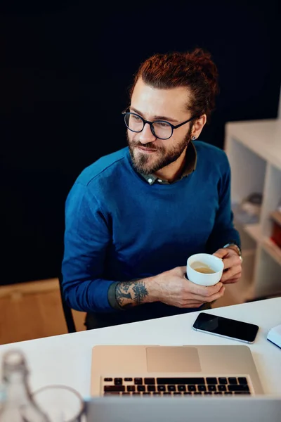 Attraktiv skäggig entreprenör sitter på sitt kontor, koppla av och dricka kaffe. — Stockfoto
