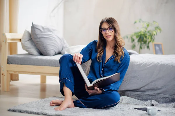 Молодая красивая брюнетка в пижаме, сидящая на полу в спальне и читающая по утрам книгу . — стоковое фото