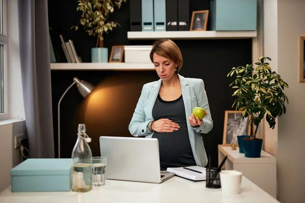 Attraktiv kaukasisk gravid affärskvinna i fyrtioårsåldern stående på kontoret, äta äpple och titta på laptop. — Stockfoto