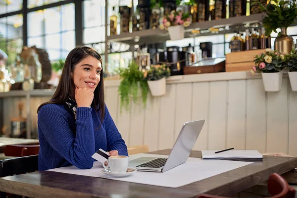 Piękna elegancka biała brunetka w swetrze siedzi w kawiarni i używa laptopa do zakupów online. W ręku jest karta kredytowa.. — Zdjęcie stockowe