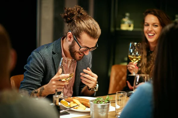 Jovem e bonito hipster caucasiano sentado com amigos no restaurante, segurando um copo de vinho e rindo . — Fotografia de Stock