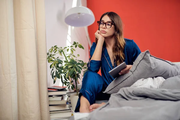 Gelangweilte niedliche kaukasische Brünette im Schlafanzug mit Brille, die morgens auf dem Bett sitzt und Tabletten in der Hand hält. — Stockfoto