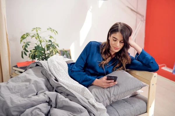 Bella bruna caucasica vestita in pigiama blu sdraiata a letto al mattino e usando lo smart phone per leggere o inviare messaggi . — Foto Stock