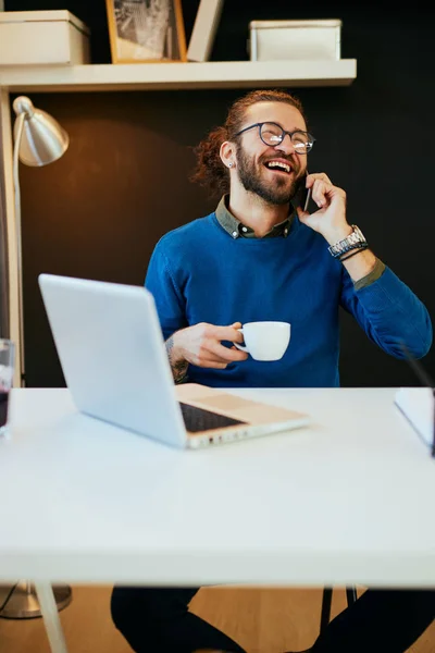 Jonge lachende blanke blogger met baard die in zijn kantoor zit, telefoneert, koffie vasthoudt en een pauze neemt van zijn werk.. — Stockfoto