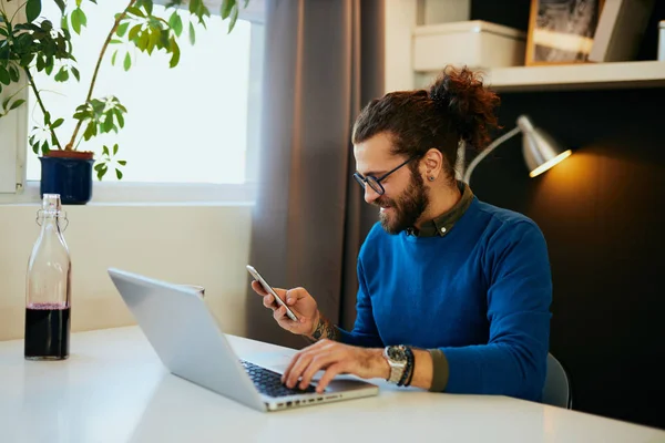 Unga leende kaukasiska entreprenör sitter i moderna kontor, hålla ena handen på laptop tangentbord och i andra smarta telefon och läsa sms. — Stockfoto