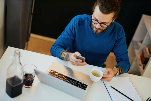 Vue aérienne d'un bel hipster caucasien barbu assis dans son bureau moderne, utilisant un téléphone intelligent pour vérifier les messages sur les médias sociaux et tenant une tasse de café . — Photo