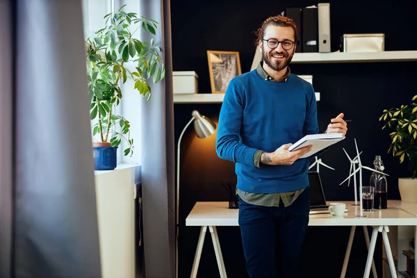 Jonge glimlachende Kaukasische innovatieve creatieve grafisch ontwerper staan in zijn kantoor en het schrijven van ideeën in notebook. — Stockfoto